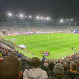 Hetman - Widok z trybun na oświetlony stadion piłkarski podczas meczu, z widocznymi kibicami i grą na murawie.