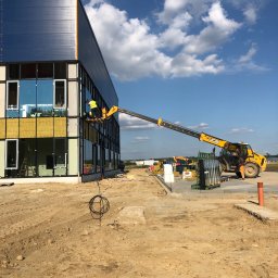 Hetman - Pracownicy na podnośniku montują elewację budynku biurowego z niebieskimi panelami i dużymi oknami, widoczne materiały budowlane i maszyny na placu budowy.