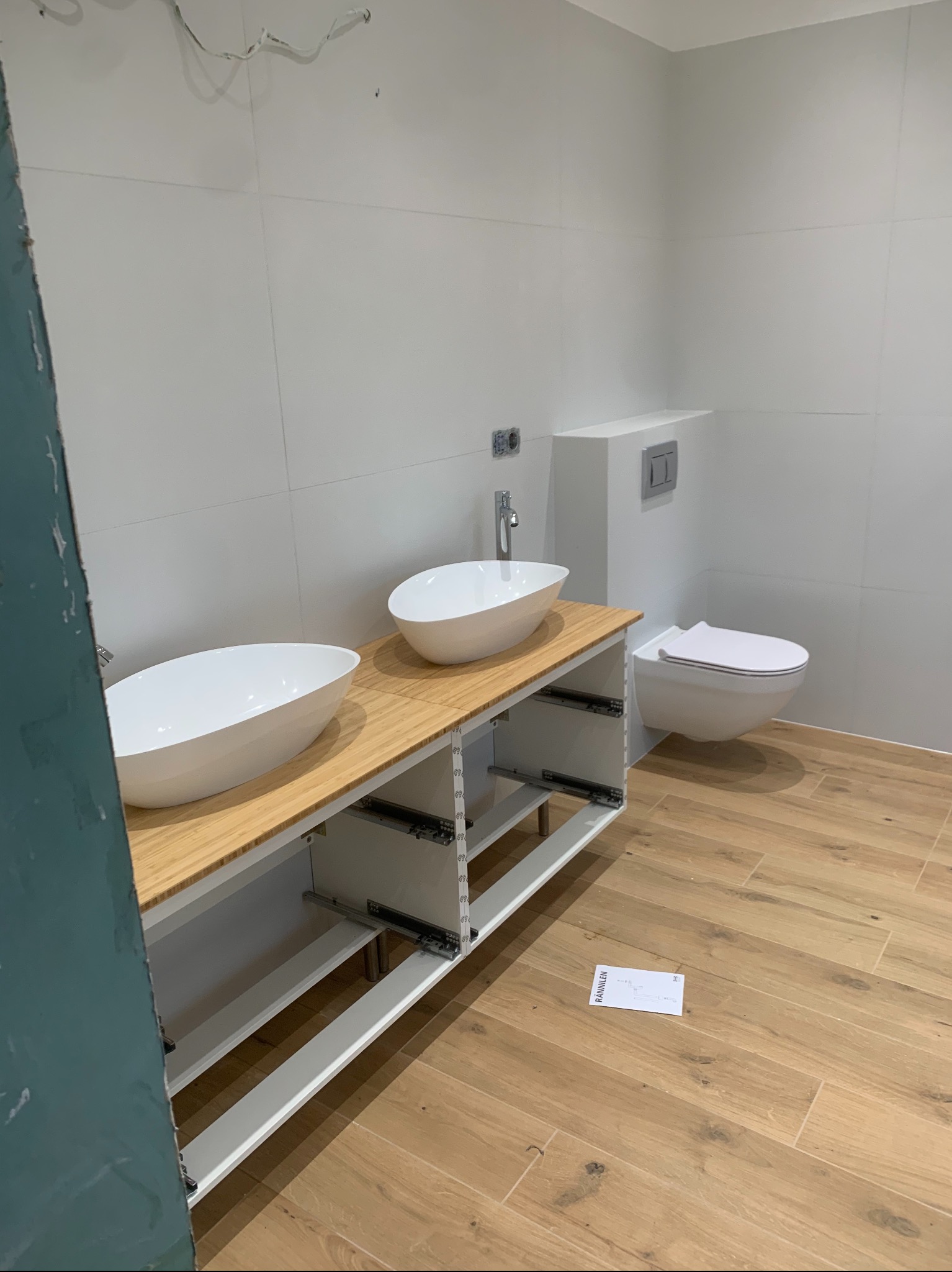 Nowoczesna łazienka w trakcie wykańczania, z dwiema owalnymi umywalkami nablatowymi na drewnianym blacie, wiszącą toaletą i płytkami imitującymi drewno na podłodze. Widoczne niedokończone elementy...