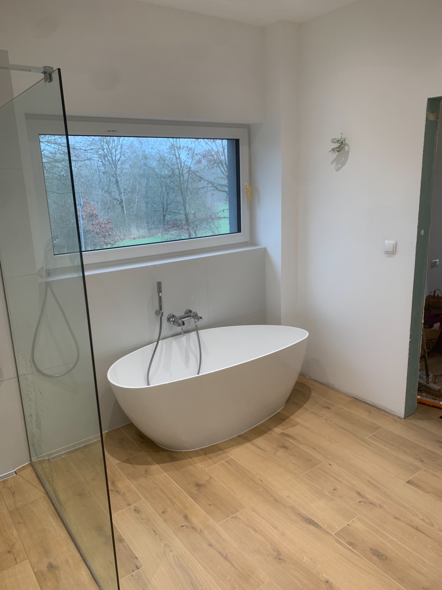 Nowoczesna, wolnostojąca wanna owalna z chromowaną armaturą w jasnej łazience z drewnopodobną podłogą i przeszkloną kabiną prysznicową; widoczne niedokończone prace wykończeniowe przy ościeżnicy...