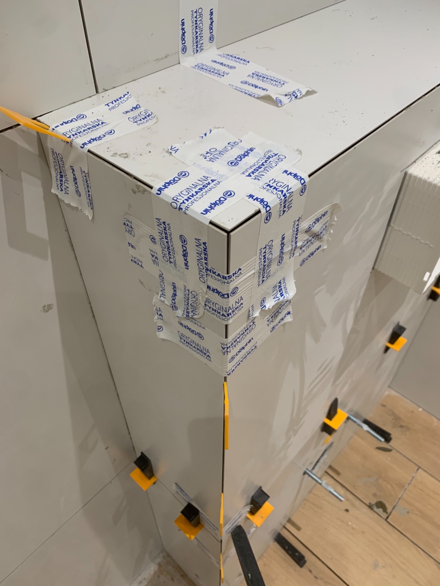 Świeżo położone, białe płytki ceramiczne na ścianie, zabezpieczone taśmą maskującą z logo producenta i stabilizowane żółtymi klipsami dystansowymi, podłoga wyłożona drewnopodobnymi panelami.