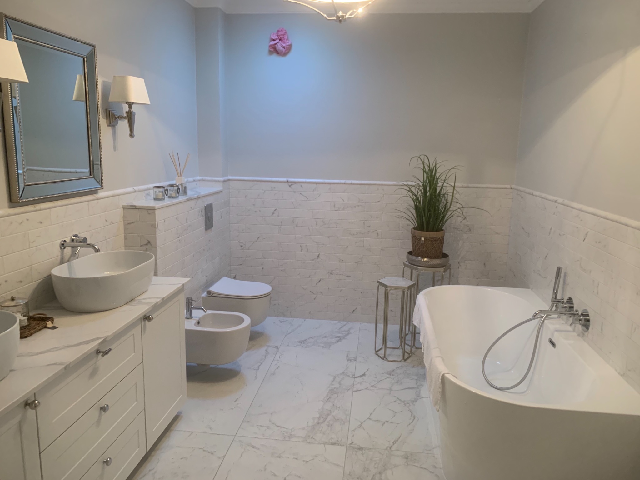 Elegancka łazienka w odcieniach bieli i szarości z marmurowymi płytkami, wolnostojącą wanną, umywalką nablatową na szafce z szufladami, bidetem i toaletą, dopełniona dekoracyjną rośliną w plecionym...