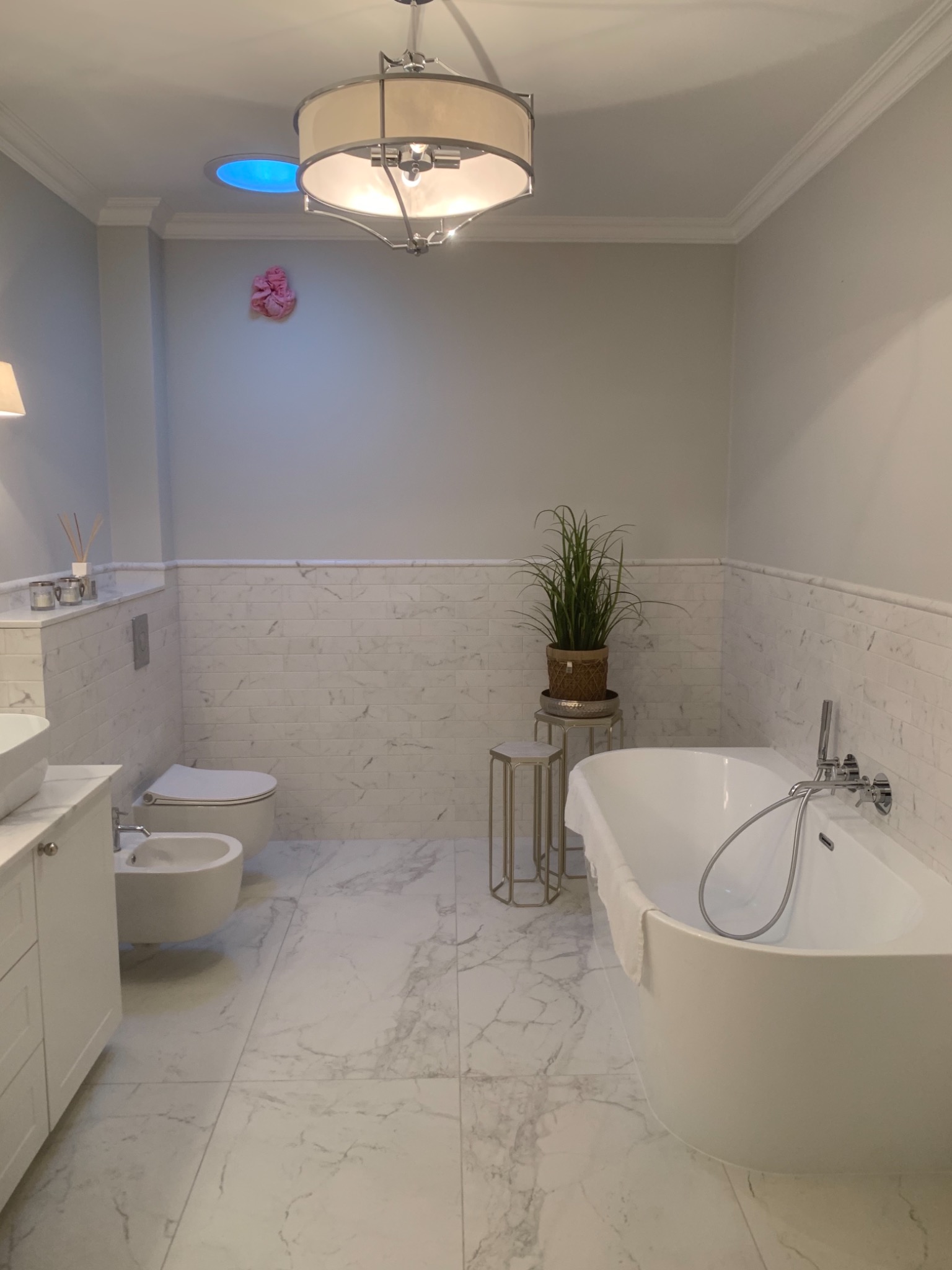Elegancka łazienka z marmurową podłogą i ścianami, białą wanną wolnostojącą z chromowaną armaturą, toaletą i bidetem, oświetlona ozdobną lampą sufitową, z rośliną doniczkową na metalowym stoliku...