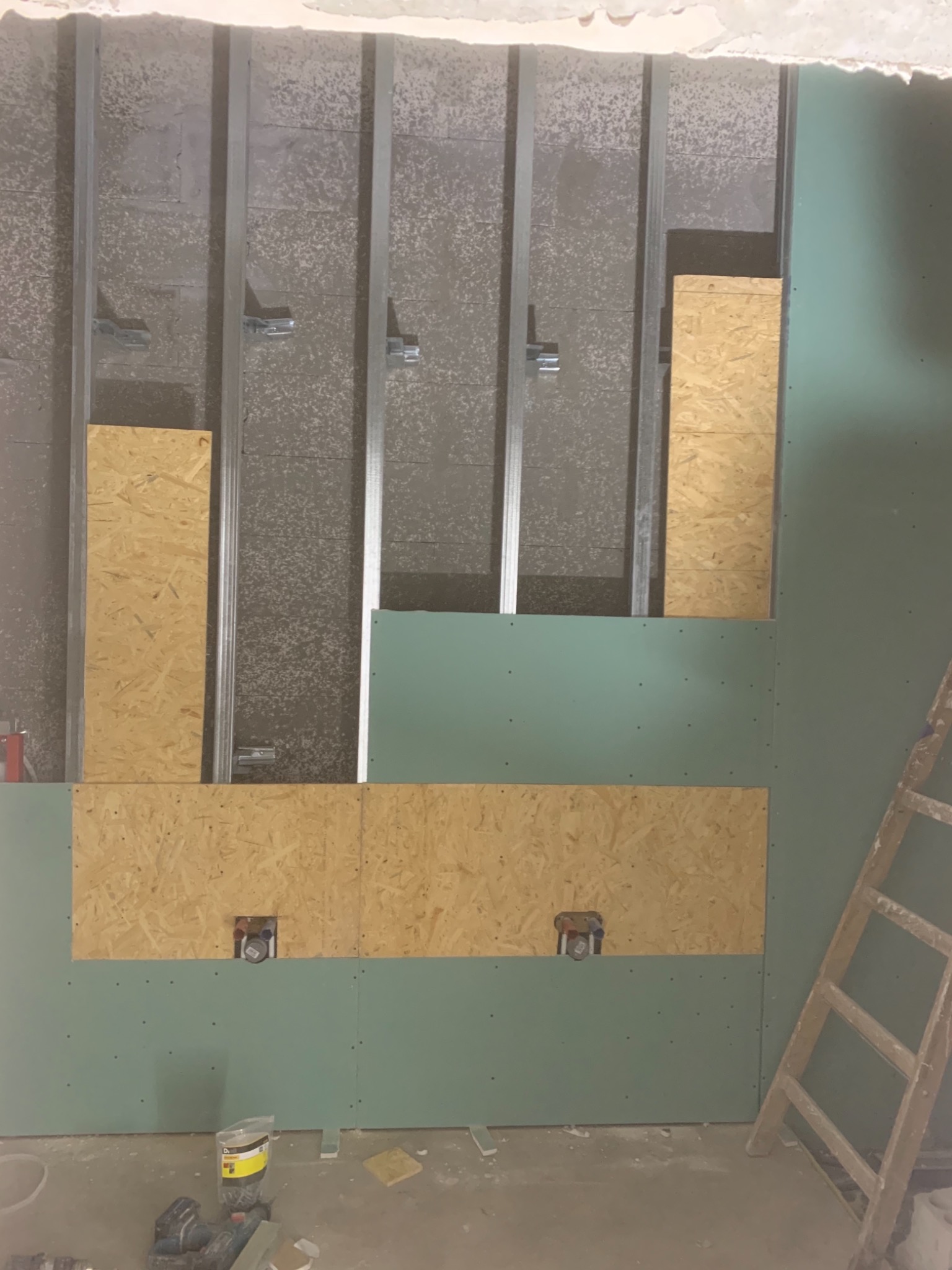 Ściana w trakcie zabudowy z płyt gipsowo-kartonowych, widoczne profile stalowe, płyty OSB i puszki elektryczne, drabina oparta o ścianę, na podłodze resztki materiałów budowlanych.