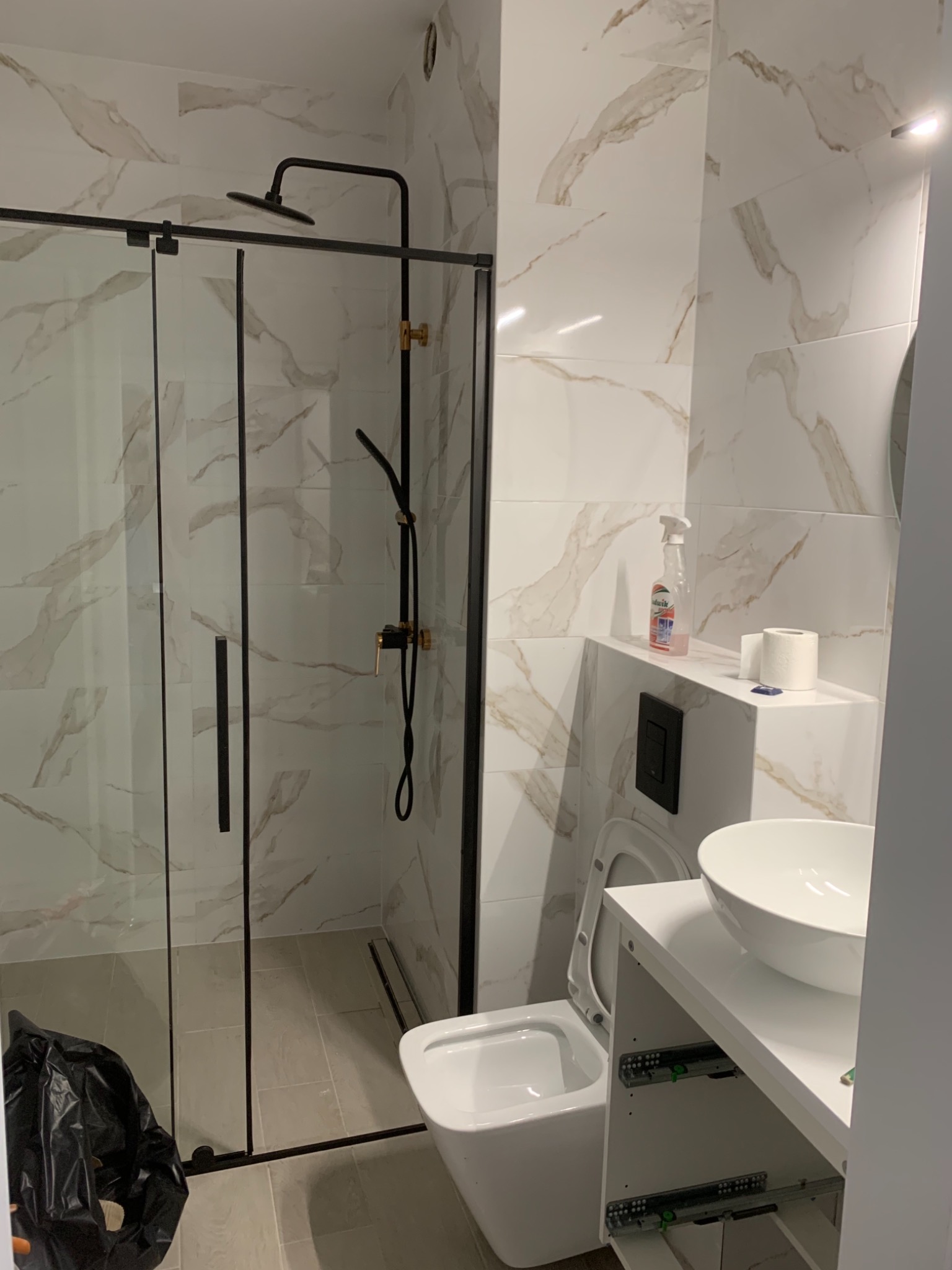 Nowoczesna łazienka z kabiną prysznicową typu walk-in z czarnymi profilami, białą umywalką nablatową i toaletą, wykończona płytkami imitującymi marmur. Worki na śmieci przy kabinie.
