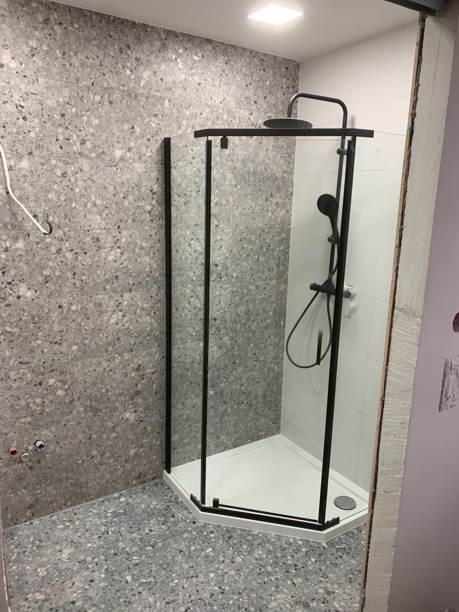 Nowoczesna kabina prysznicowa z czarną ramą, biały brodzik, ściany i podłoga wyłożone szarą mozaiką, widoczne rury instalacji wodnej.