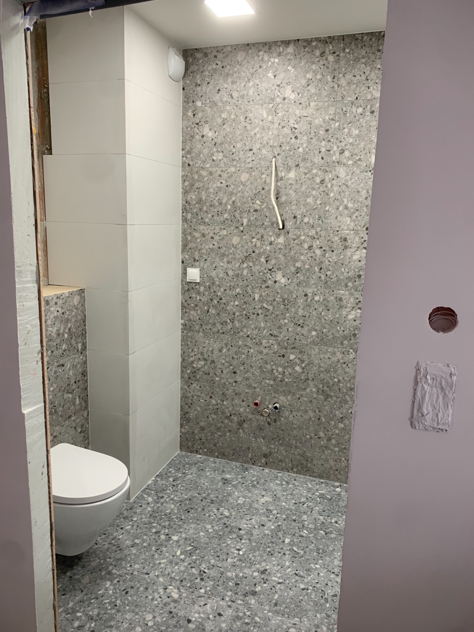 Łazienka w trakcie wykańczania: biała toaleta, szare płytki lastryko na ścianach i podłodze, widoczne wystające kable i rury, ściana pomalowana na fioletowo z niedokończonymi poprawkami.