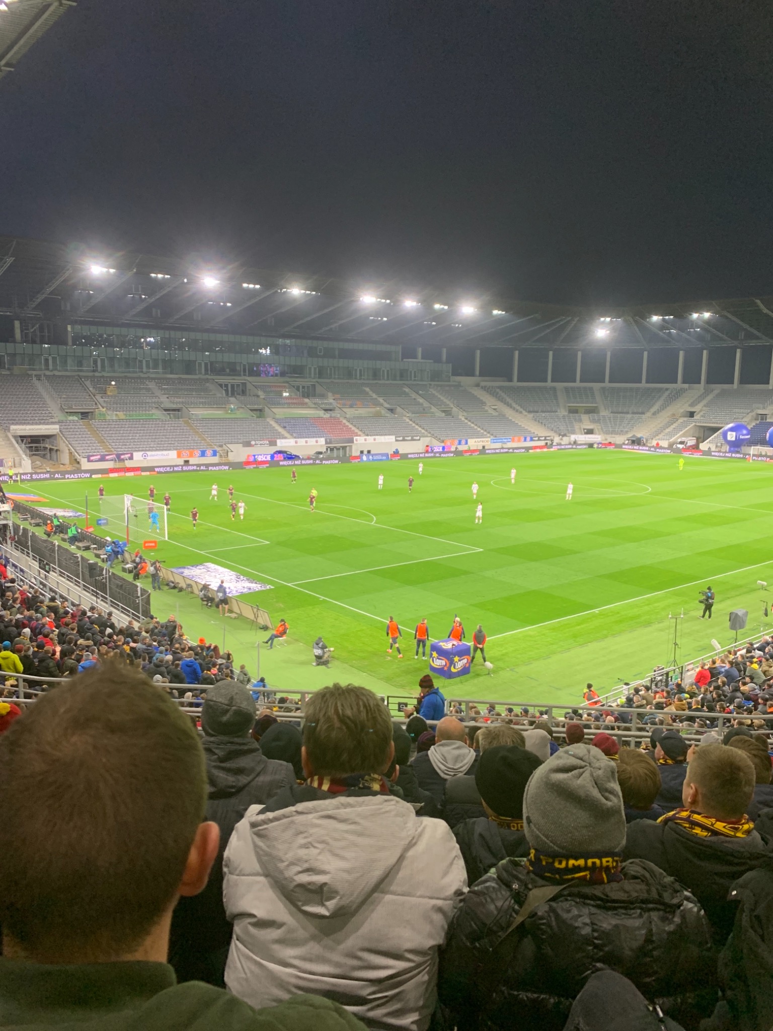Widok z trybun na oświetlony stadion piłkarski podczas meczu, z widocznymi kibicami i grą na murawie.