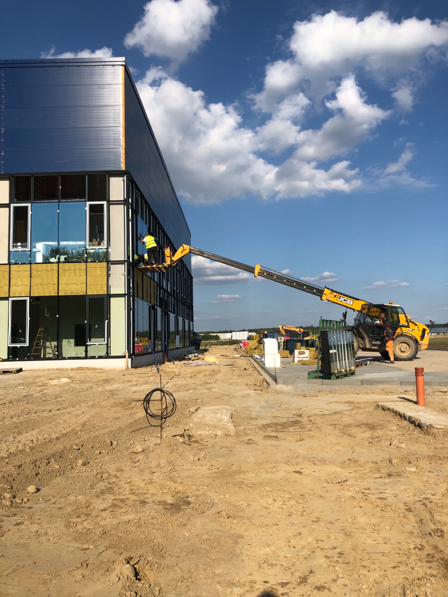 Pracownicy na podnośniku montują elewację budynku biurowego z niebieskimi panelami i dużymi oknami, widoczne materiały budowlane i maszyny na placu budowy.