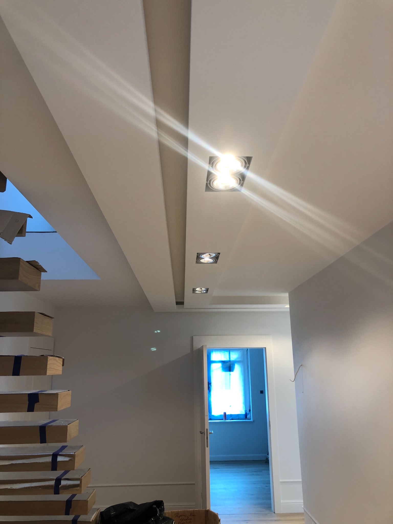 Nowoczesne wykończenie sufitu z oświetleniem punktowym i schody z zabezpieczonymi stopniami w jasnym wnętrzu, widok na otwarte drzwi i okno z niebieską zasłoną w tle.