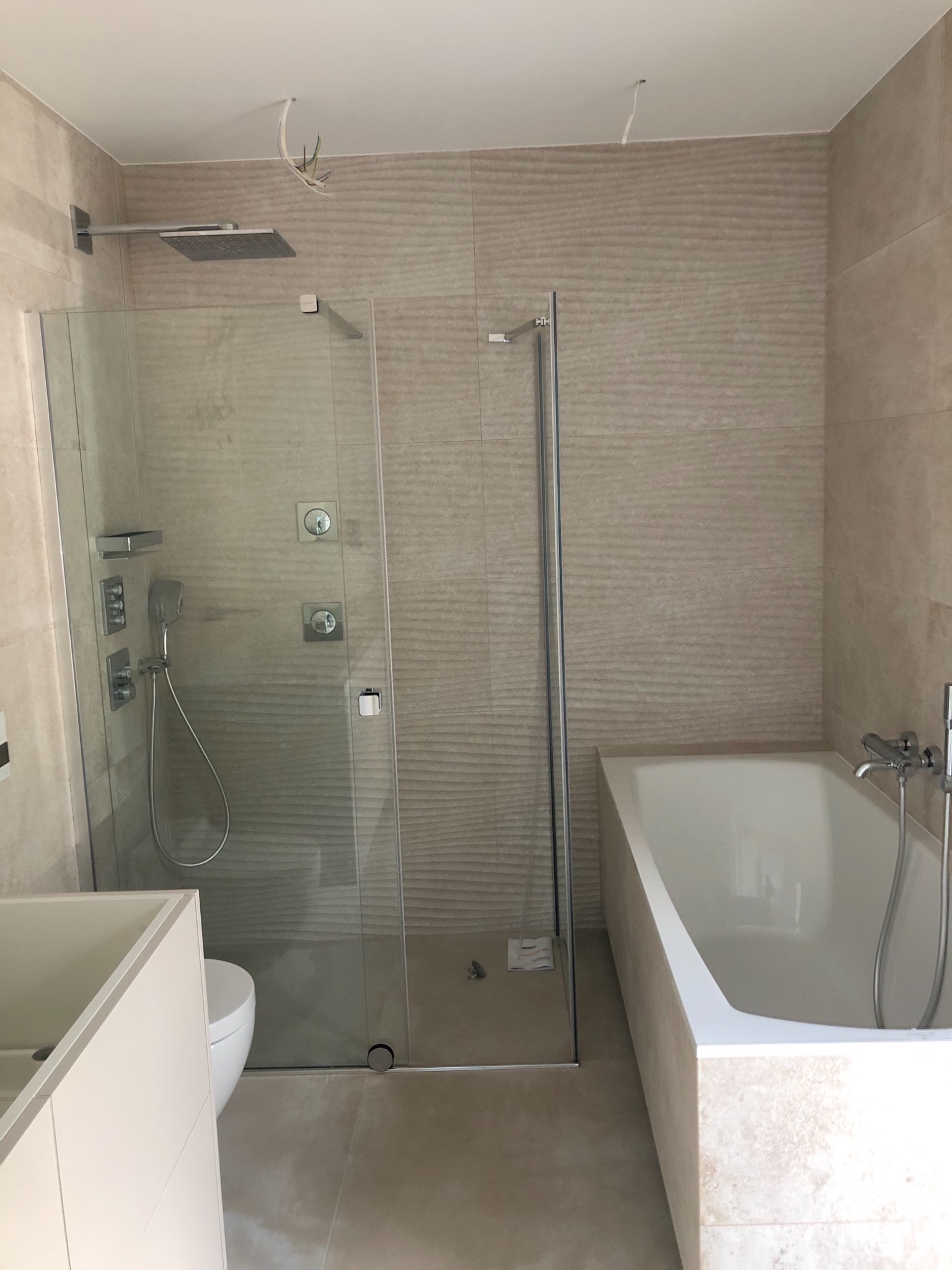 Nowoczesna łazienka z kabiną prysznicową typu walk-in, wanną obudowaną płytkami i beżowymi płytkami ściennymi o falistej strukturze. Widoczne wystające kable z sufitu.