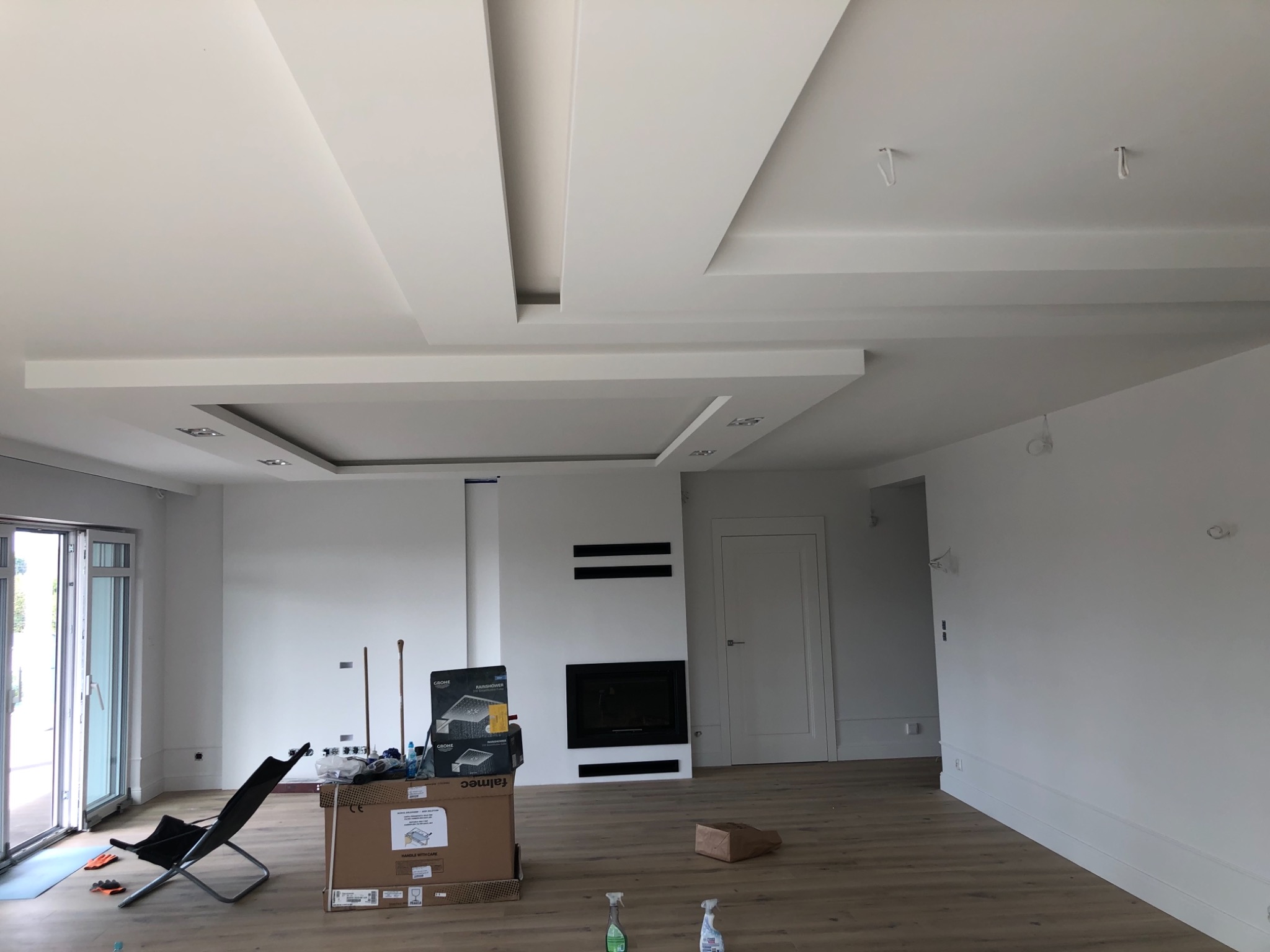 Jasne wnętrze w trakcie wykończenia, z dekoracyjnym podwieszanym sufitem, widoczne kartony z armaturą, krzesło i środki czystości na podłodze.