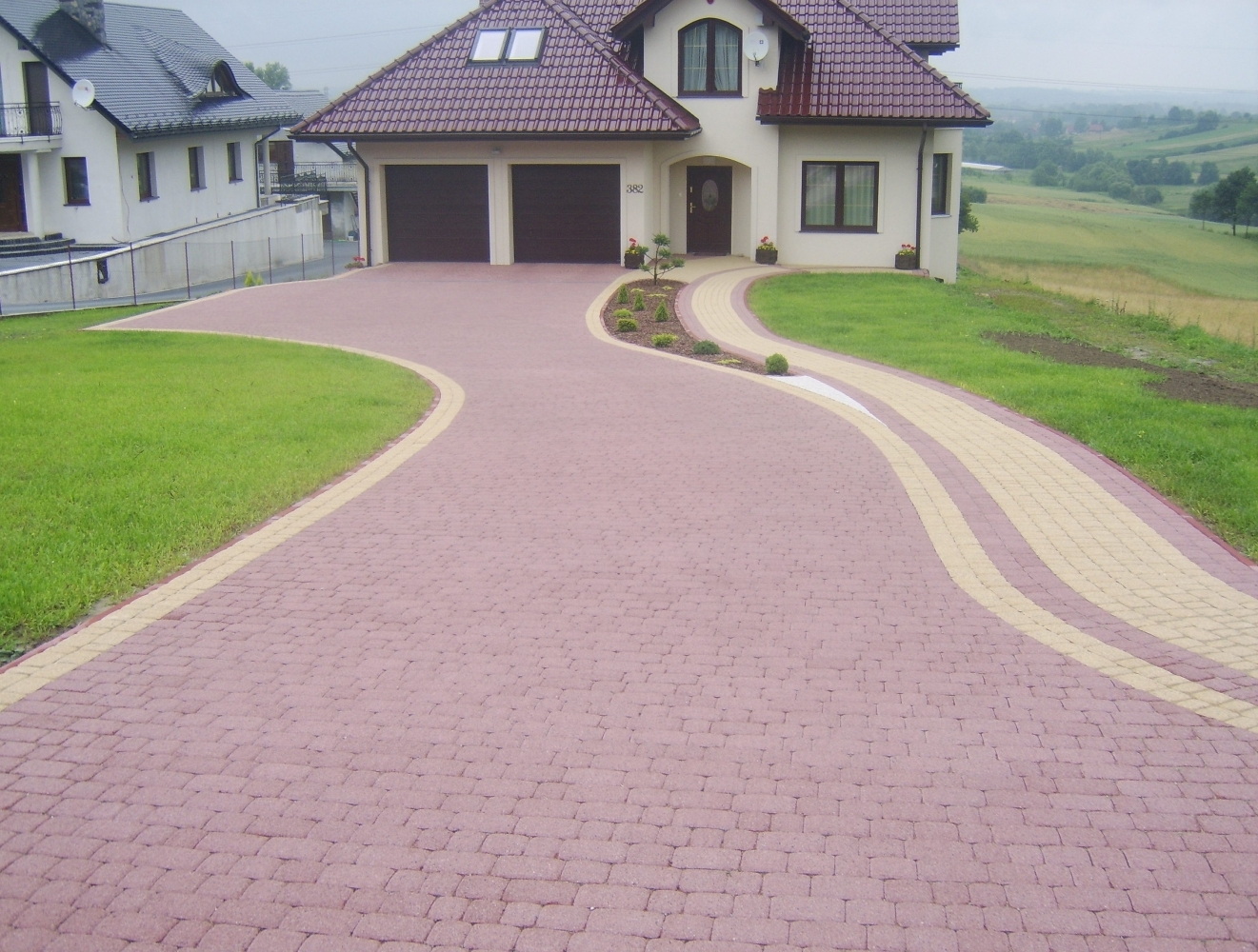 Szeroki, kręty podjazd z różowej kostki brukowej z beżowymi obrzeżami i pasami, prowadzący do dwustanowiskowego garażu w nowoczesnym domu z brązowym dachem.