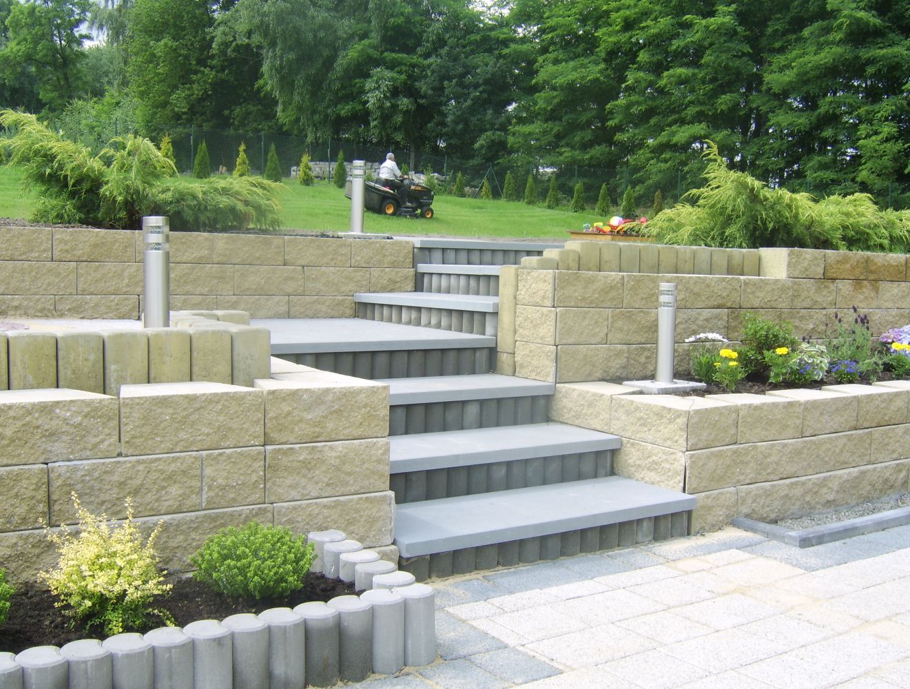 Schody z kostki betonowej prowadzące na trawnik, z widocznym pracownikiem koszącym trawę kosiarką samojezdną w tle, obok schodów rabaty kwiatowe i oświetlenie.