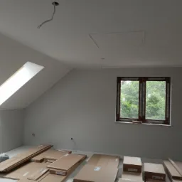 Poddasze w trakcie remontu, z oknem dachowym i zwykłym oknem z widokiem na drzewa, przygotowane panele podłogowe w kartonach i folii, wystające kable elektryczne, ściany pomalowane na szaro.