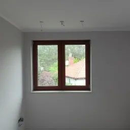 Pomalowane na szaro wnętrze pokoju na poddaszu z brązowym oknem, widoczne wystające przewody oświetleniowe z sufitu i puszki elektryczne w ścianie.