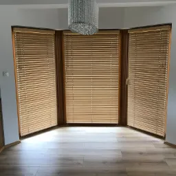 Jasne, drewniane żaluzje poziome zamontowane w oknach wykuszowych, z widocznym ozdobnym żyrandolem kryształowym i panelami podłogowymi w odcieniach szarości.