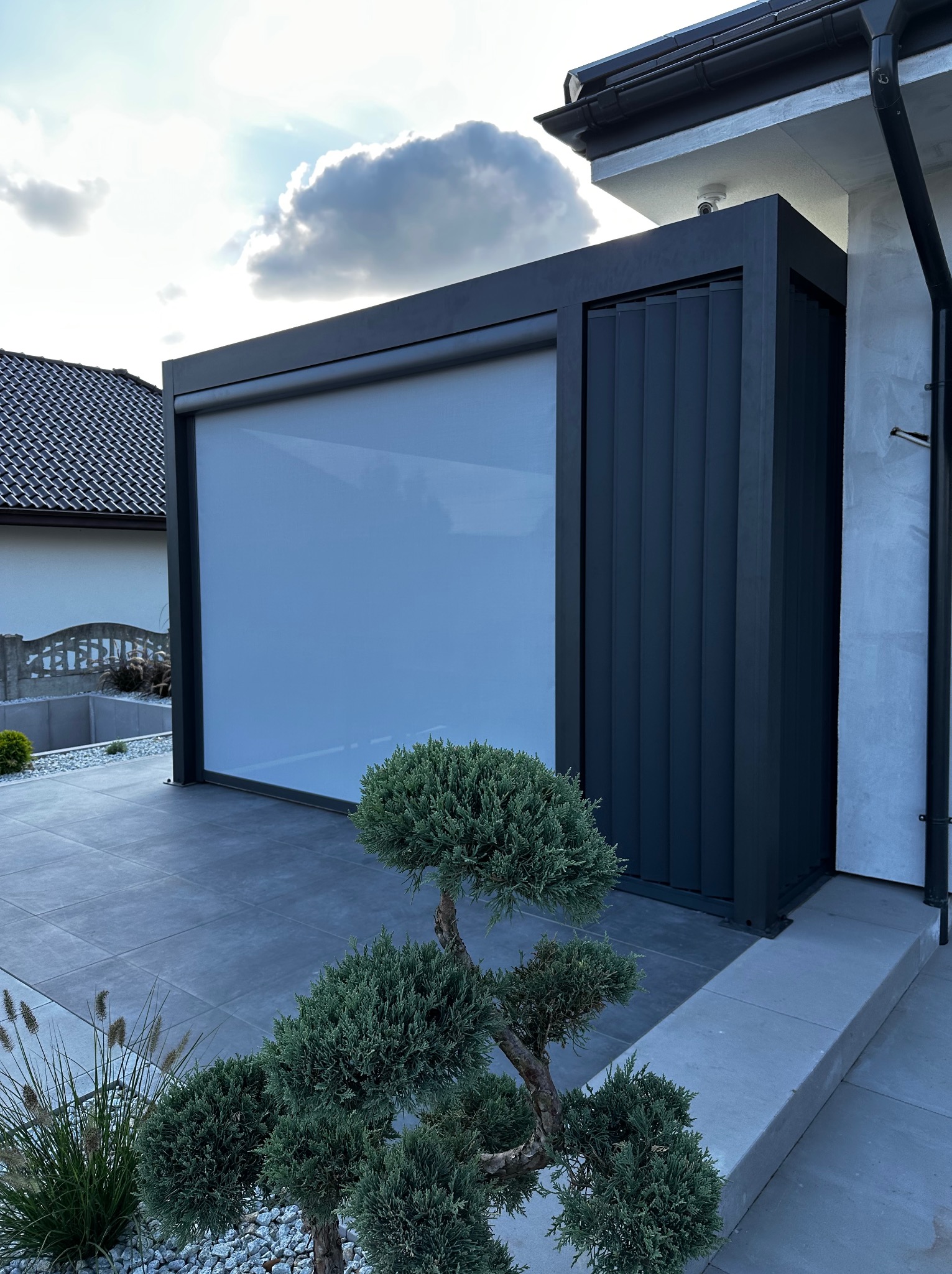 Nowoczesna pergola lamelowa z pionowymi shuttersami i roletą screen zip, widok z zewnątrz, zieleń ozdobna na pierwszym planie, szara kostka brukowa.