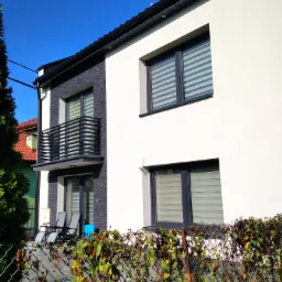 Dwukondygnacyjny dom z białą elewacją i fragmentem ściany pokrytym szarą okładziną imitującą kamień; balkon z czarną balustradą, widoczne okna z żaluzjami poziomymi, na tarasie leżaki i niebieskie...