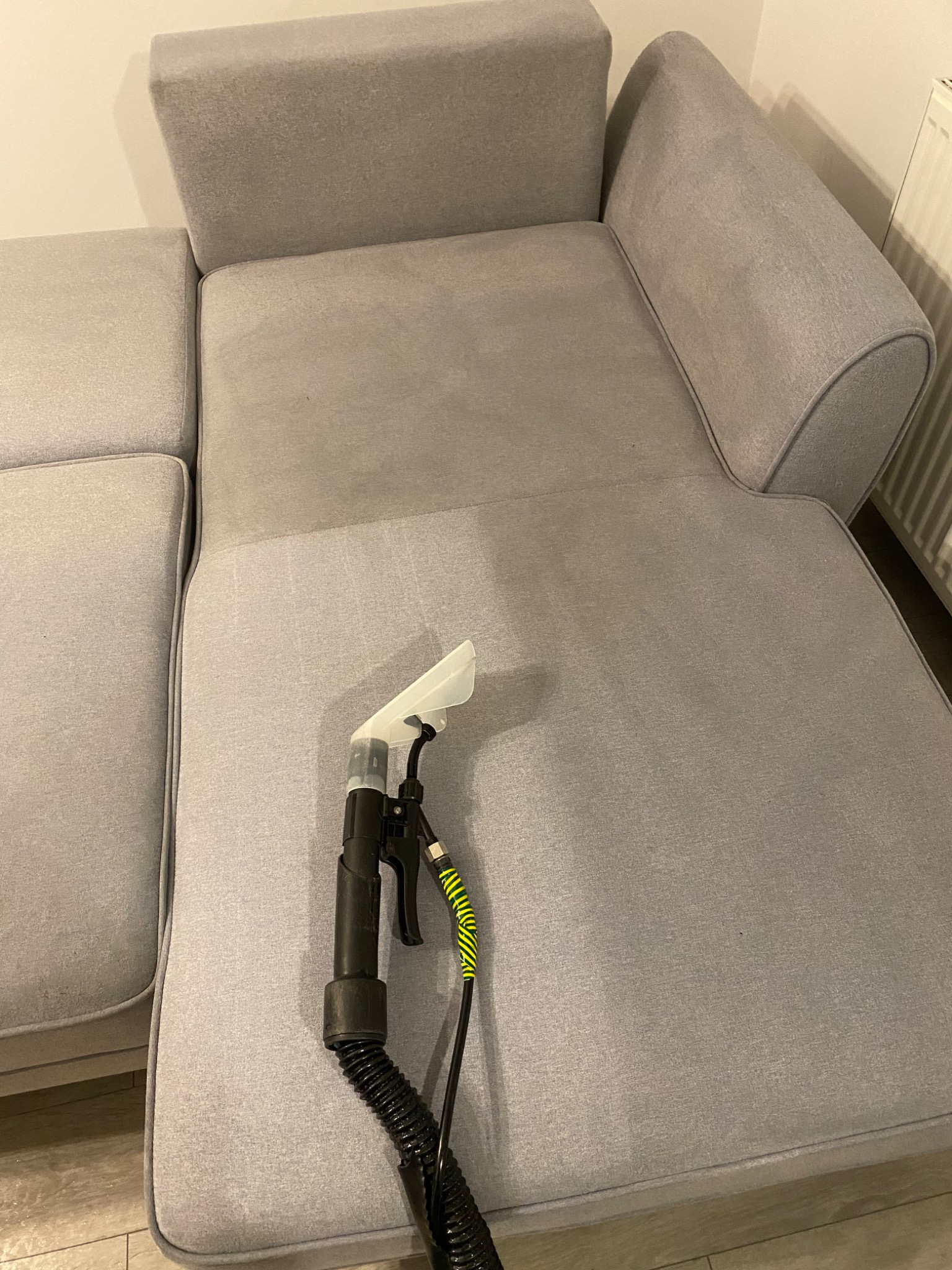 Czyszczenie jasnoszarej sofy za pomocą ekstraktora z przezroczystą dyszą; widoczna różnica w odcieniu po czyszczeniu.