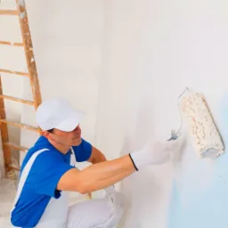 Malarz w białym kombinezonie i niebieskiej koszulce, z wałkiem malarskim w dłoni, maluje ścianę na jasny błękit, drewniana drabina oparta o ścianę w tle.