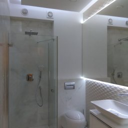 J&S Usługi remontowo budowlane - Nowoczesna łazienka z kabiną prysznicową ze szklanymi drzwiami, umywalką nablatową, podwieszaną miską WC i podświetleniem LED na suficie oraz dekoracyjnej ścianie.