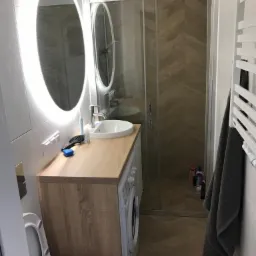 Nowoczesna łazienka z owalnym lustrem podświetlanym LED, kabiną prysznicową z wzorem jodełki na ścianie, pralką zabudowaną pod blatem z umywalką oraz grzejnikiem drabinkowym z szarym ręcznikiem.