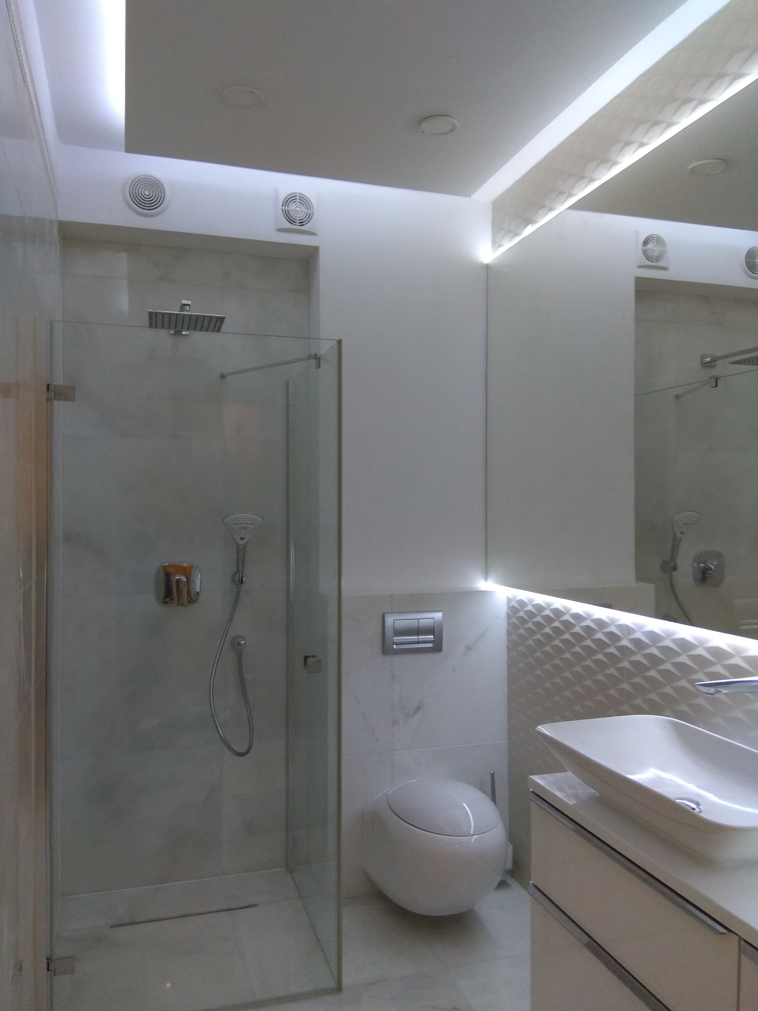 Nowoczesna łazienka z kabiną prysznicową ze szklanymi drzwiami, umywalką nablatową, podwieszaną miską WC i podświetleniem LED na suficie oraz dekoracyjnej ścianie.