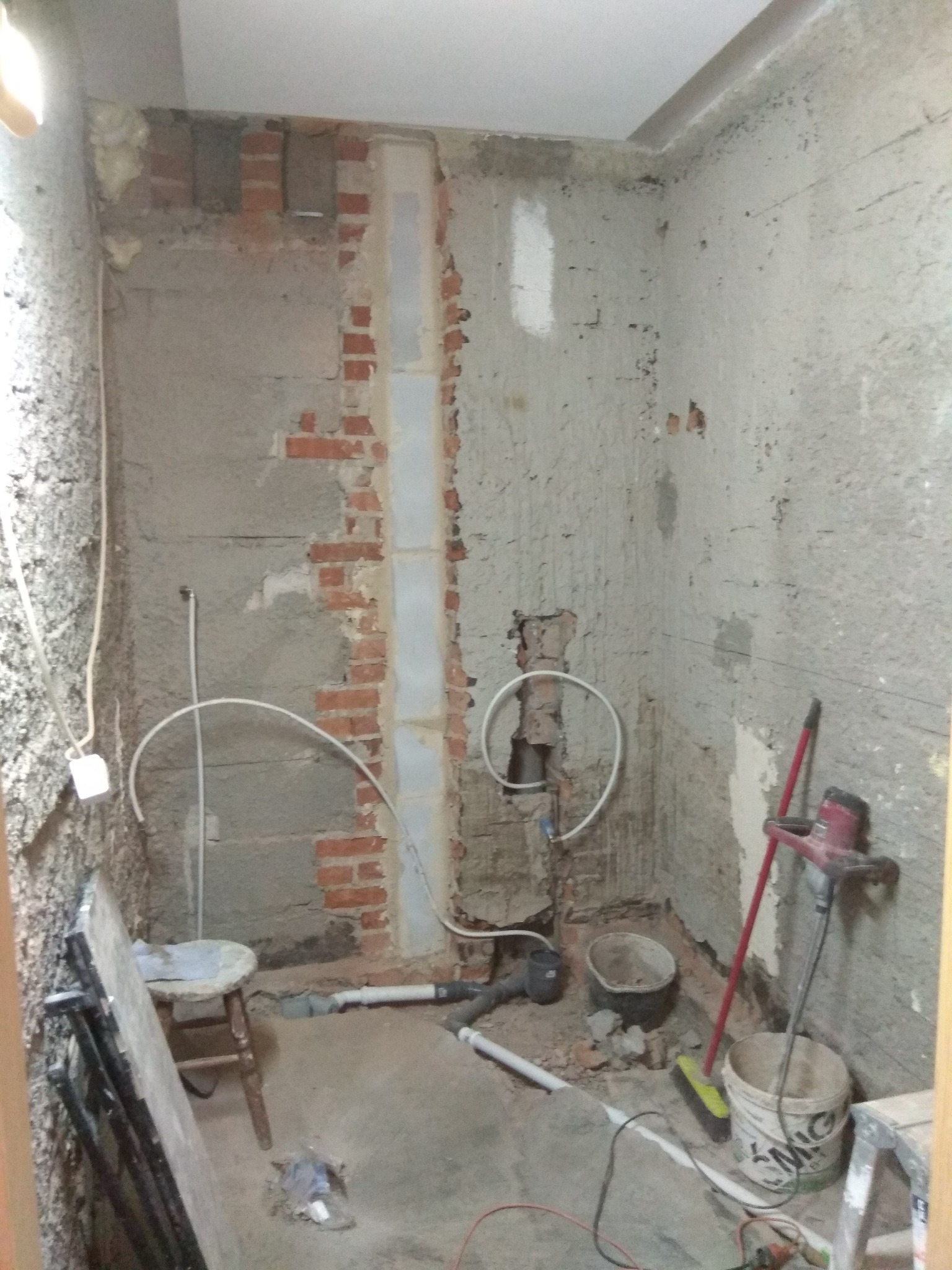 Surowy stan wnętrza łazienki w trakcie prac: odsłonięta cegła, wystające rury, stare tynki, widoczne narzędzia i wiaderka.