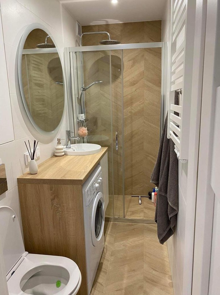 Nowoczesna łazienka z kabiną prysznicową, umywalką nablatową na drewnianym blacie, pralką w zabudowie i płytkami w jodełkę na podłodze i pod prysznicem. Na blacie dyfuzor zapachowy z czarnymi...