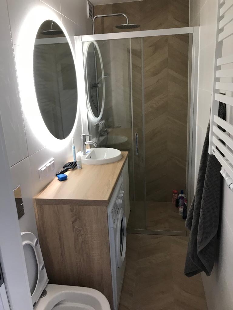 Nowoczesna łazienka z owalnym lustrem podświetlanym LED, kabiną prysznicową z wzorem jodełki na ścianie, pralką zabudowaną pod blatem z umywalką oraz grzejnikiem drabinkowym z szarym ręcznikiem.