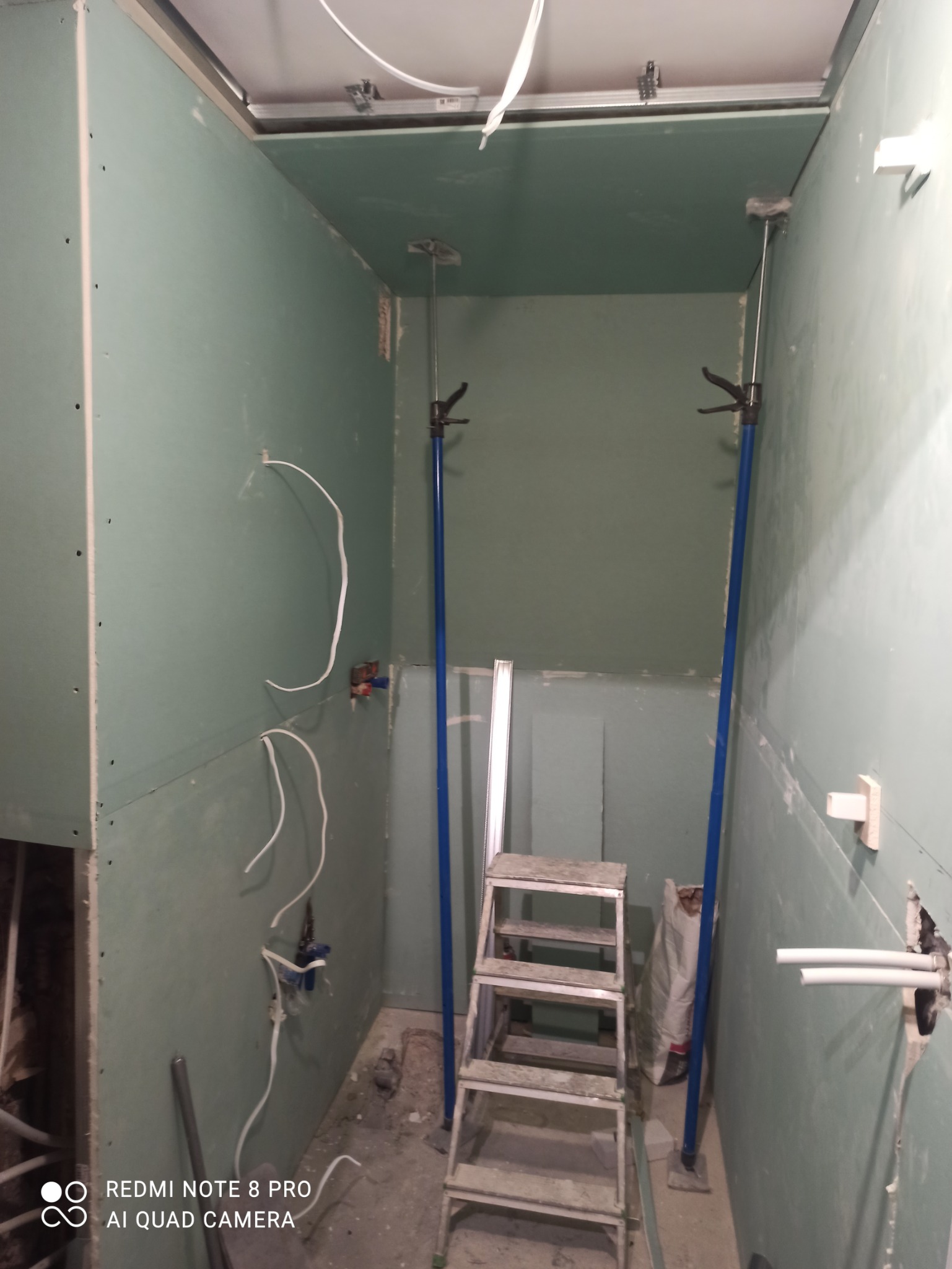 Wnętrze w trakcie prac przygotowawczych: ściany i sufit obłożone płytami gipsowo-kartonowymi, widoczne instalacje elektryczne, drabina aluminiowa i podpory teleskopowe.