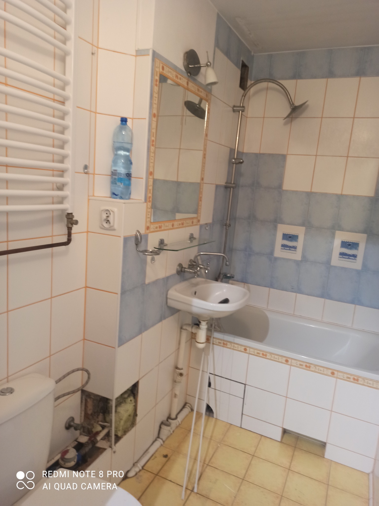 Ujęcie wnętrza łazienki z widoczną toaletą, umywalką na metalowym stelażu, wanną i grzejnikiem drabinkowym. W ścianie widoczny otwór instalacyjny.