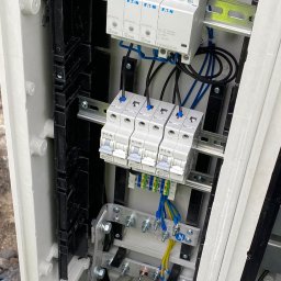 Elektroinstalacje Łukasz Janik - Elektryk w rękawicach ochronnych MaxiFoam podłącza przewód do szyny wyrównawczej wewnątrz skrzynki elektrycznej z zamontowanymi bezpiecznikami i ogranicznikiem przepięć marki Eaton.