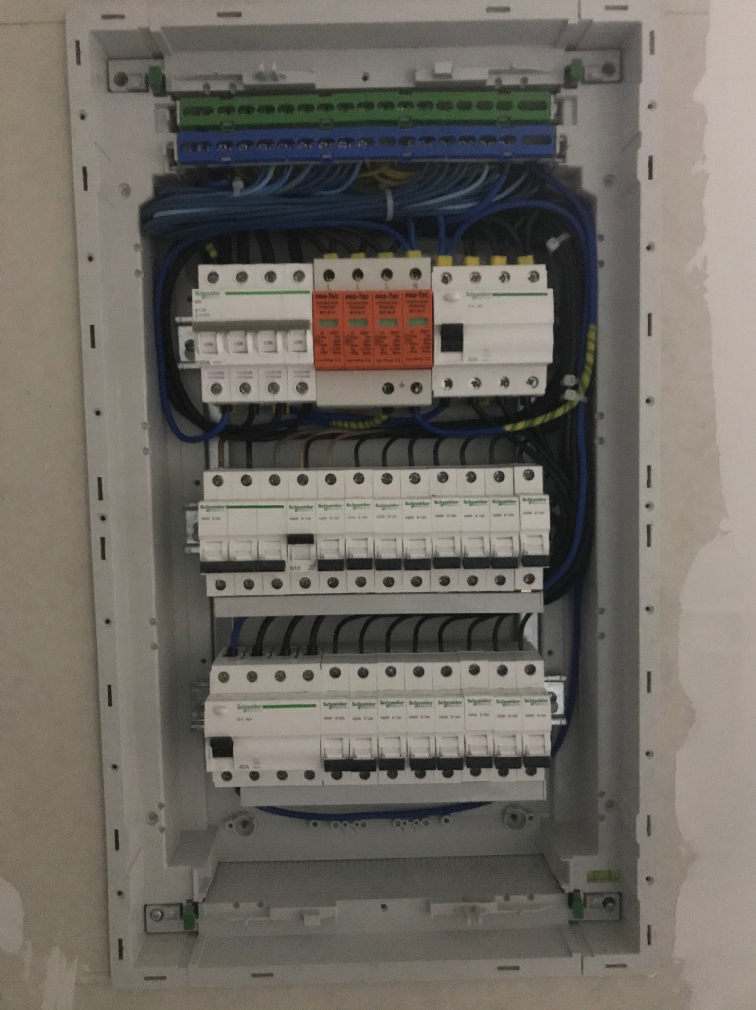 Otwarta skrzynka elektryczna w ścianie, widoczne bezpieczniki, przewody i listwy zaciskowe, ściana wokół skrzynki nieotynkowana.