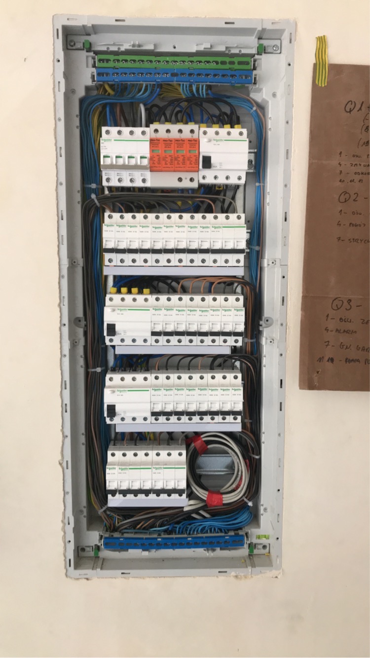 Rozdzielnica elektryczna wnękowa z widocznymi zabezpieczeniami, przewodami i listwami zaciskowymi, obok kartka z odręcznymi notatkami przyklejona taśmą.