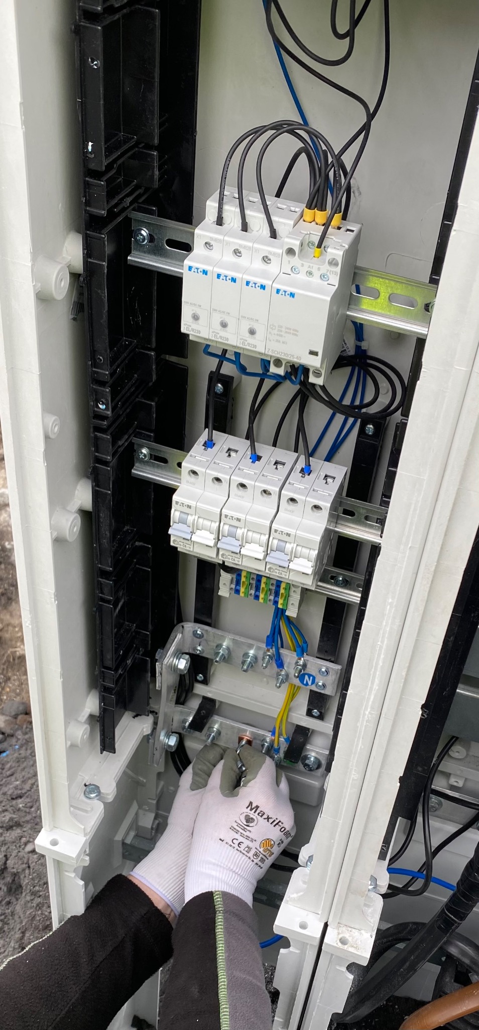 Elektryk w rękawicach ochronnych MaxiFoam podłącza przewód do szyny wyrównawczej wewnątrz skrzynki elektrycznej z zamontowanymi bezpiecznikami i ogranicznikiem przepięć marki Eaton.