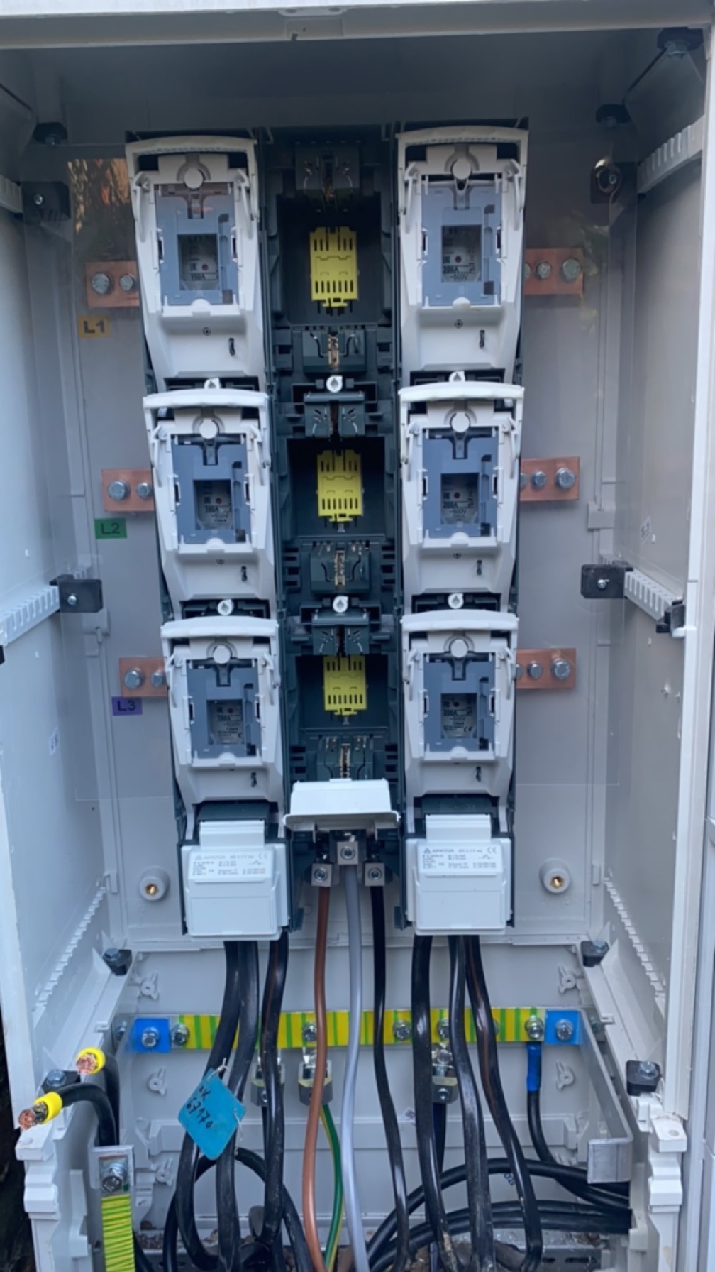 Otwarta skrzynka elektryczna z zainstalowanymi bezpiecznikami, okablowaniem i szynami zbiorczymi; widoczne oznaczenia L1, L2, L3 oraz niebieska etykieta z numerem seryjnym.