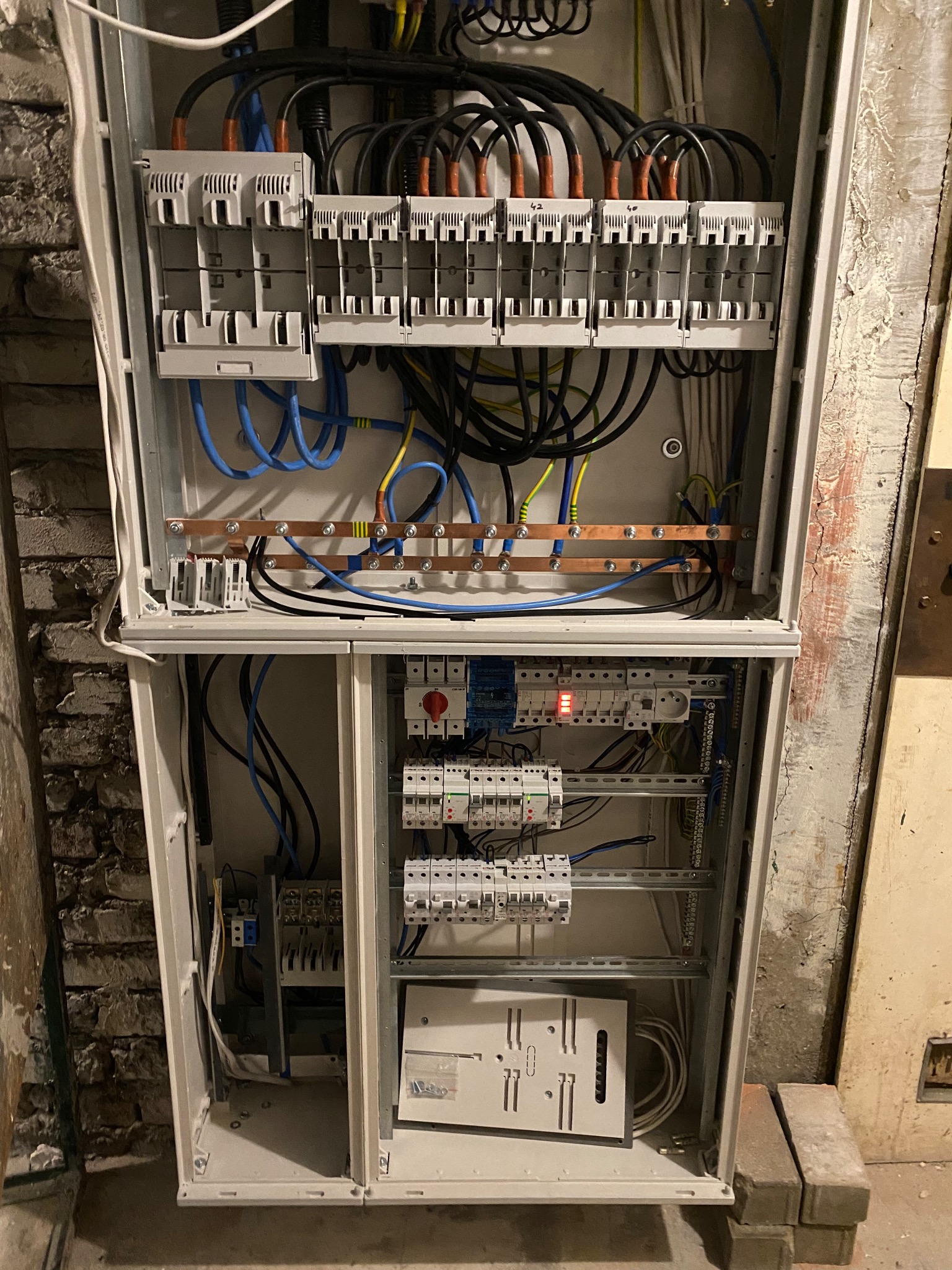 Otwarta skrzynka rozdzielcza z widocznymi bezpiecznikami, przewodami i szynami, umieszczona na tle ściany z elementami cegieł i farby.