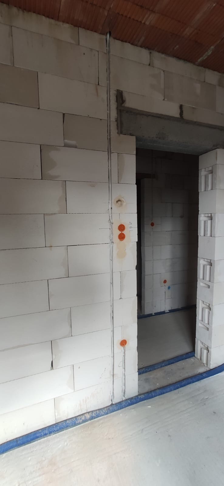 Surowy stan budynku z widocznymi przewodami elektrycznymi w ścianie z betonu komórkowego, zabezpieczonymi pomarańczowymi puszkami instalacyjnymi i niebieską rurą karbowaną przy podłodze.
