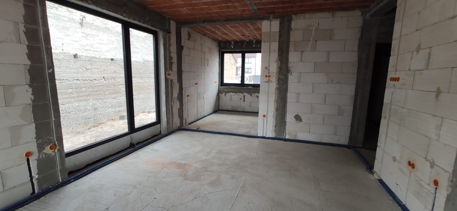 Surowy stan wnętrza z widocznymi instalacjami elektrycznymi w rurkach ochronnych na ścianach z betonu komórkowego, otwory okienne z czarnymi ramami, sufit wykończony cegłą dziurawką.