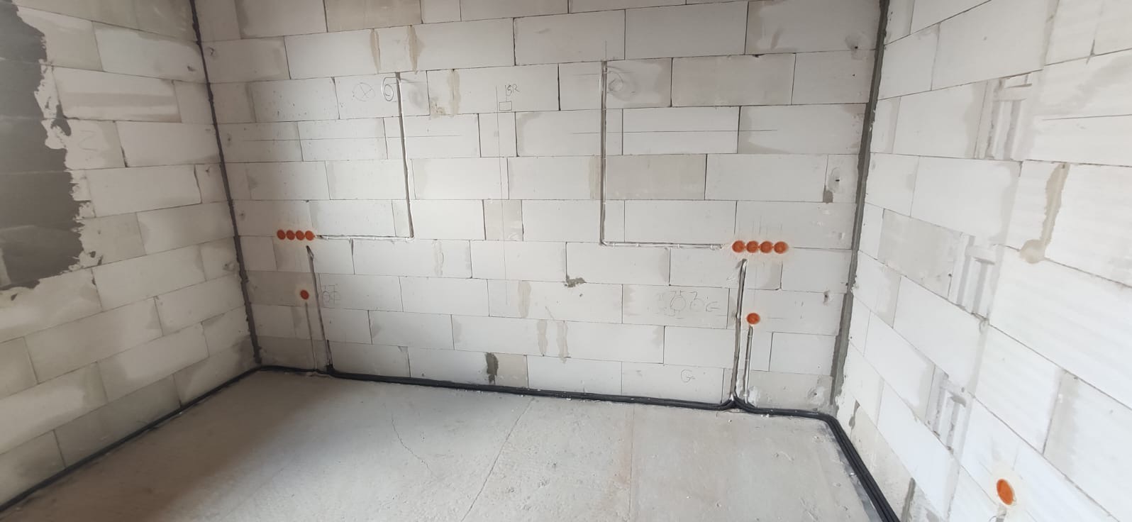 Surowe wnętrze z białych bloczków, widoczne bruzdy pod kable elektryczne i pomarańczowe puszki instalacyjne na ścianach, czarne kable prowadzone wzdłuż podłogi.