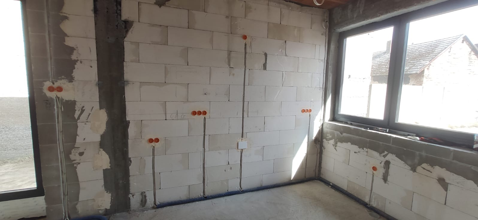 Surowe wnętrze w trakcie budowy z widocznymi przewodami elektrycznymi w ścianach z betonu komórkowego, pomarańczowe puszki instalacyjne i niebieska rurka karbowana przy podłodze.