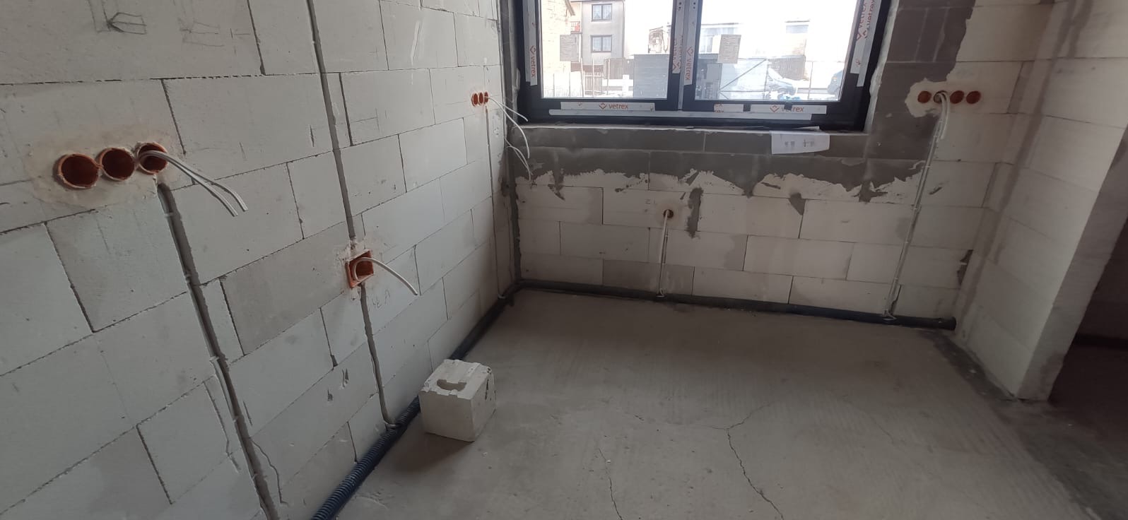 Surowy stan wnętrza z widocznymi puszkami instalacyjnymi, przewodami elektrycznymi w bruzdach w ścianach z betonu komórkowego, oraz rurą instalacyjną przy podłodze i oknem z widokiem na budynek...