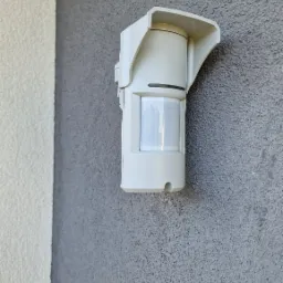 Zewnętrzny czujnik ruchu alarmowego zamontowany na szarej, chropowatej ścianie budynku, widoczne łączenie z białą elewacją.