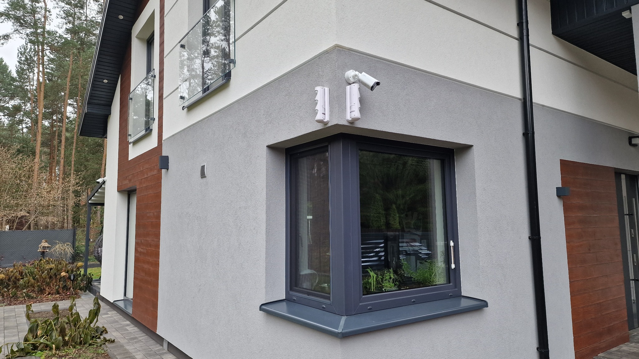 Zewnętrzny monitoring domu: kamera IP na elewacji nowoczesnego domu z szarą stolarką okienną i dekoracyjną okładziną drewnopodobną.