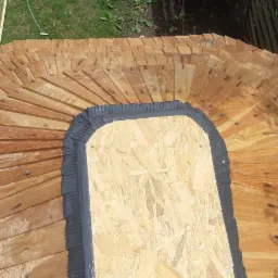 Fragment dachu krytego drewnianymi gontami w kształcie łuski, z widocznym wycięciem i zabezpieczeniem z czarnej taśmy uszczelniającej, w tle trawnik i elementy konstrukcyjne.