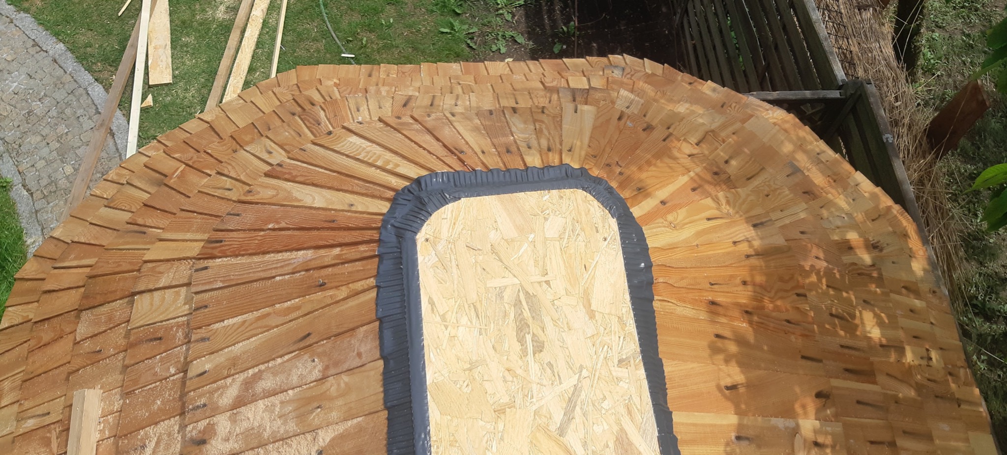 Fragment dachu krytego drewnianymi gontami w kształcie łuski, z widocznym wycięciem i zabezpieczeniem z czarnej taśmy uszczelniającej, w tle trawnik i elementy konstrukcyjne.