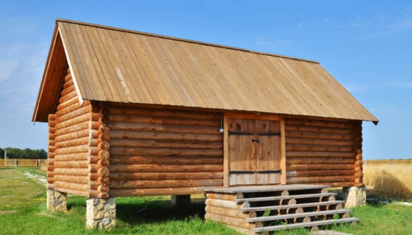 Drewniany budynek z bali z dwuspadowym dachem z desek, uniesiony na betonowych fundamentach, z drewnianymi schodami wejściowymi, na tle błękitnego nieba i zielonego trawnika.