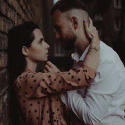 Red Dot Kamil Matuszewski - Para zakochanych, kobieta w sukience w grochy oparta o ceglaną ścianę, mężczyzna w białej koszuli obejmuje ją, wzajemne spojrzenia, tatuaż na ramieniu mężczyzny.